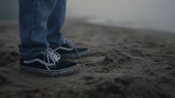 Closeup stylish guy in sneakers standing on sandy beach. Unrecognizable man legs dancing on sea shore. Male dancer stepping in music rhythm. Man moving feet. Hipster wearing jeans and casual shoes