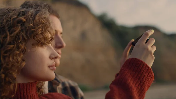 在日出海滩用智能手机拍照的轻松自在的恋人的画像 侧视图聚焦夫妇面对享受日落景观 梦想中的朋友站在海滨 用手机写博客的女孩 — 图库照片
