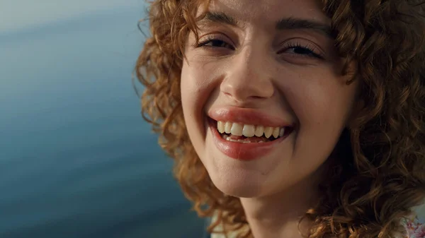 Closeup of attractive face happy young girl in front blue ocean. Cheerful curly lady smiling looking camera on sunshine. Portrait of carefree pretty woman on sunny seacoast. Natural beauty concept.