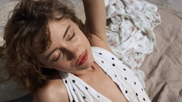 Seductive attractive girl in polka dot white swimsuit putting face under sunlight close up. Portrait of sexy curly woman sunbathing on beach sand. Beautiful lady enjoying summer sunshine on vacation.