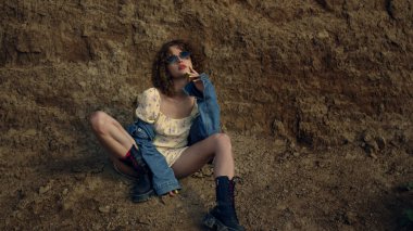 Kum mevsiminde rahat bir şekilde oturan kendinden emin seksi kadın. Doğada poz veren havalı güneş gözlüğü takan çekici bir genç kız. Çekici bir model, kameraya dokunuyor. Moda konsepti.