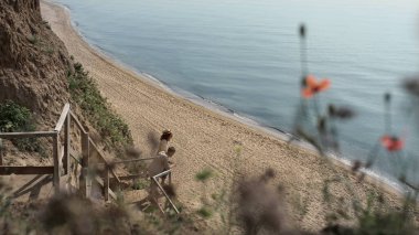 Rahat aşıklar deniz manzarasını uzaktan kumlu sahilde seyrediyorlar. Genç çift okyanusta tatil yaparken birlikte dinleniyor. Güzel bir çift, güneşli bahar zamanının tadını çıkarır..