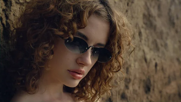 Pretty face young model looking camera confidently in sunglasses. Portrait of attractive sexy woman leaning on sand hill outdoors. Curly serious girl posing standing on seacoast alone. Relax on nature