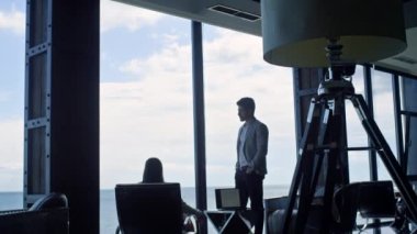 Business couple silhouettes thinking at marine view. Two unknown coworkers discussing business deal at panorama window. Unrecognized partners having conversation in dark interior conference room 