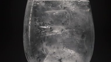 Buz blokları soğuk su bardağı yakın plan. Şarap kadehinde buzlu alkollü cin soda, ağır çekimde ışıldıyor. Siyah arka plan oluşturan serin, berrak içecek. Tonik kavramı tazeleniyor