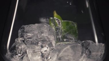 Nane garnitürlü buz kokteyli yakın plan. Yeşil yapraklar buz blokları makro siyah arka plan süper yavaş çekim ile soğuk cama düşer. Taze malzeme konseptiyle parti içeceği hazırlıyorum.