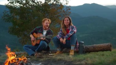 Two campers rest outdoors in mountains view. Happy couple spend vacation outdoors closeup. Smiling tourists enjoy play guitar music by campfire. Romantic family have fun at holiday. Youth concept.