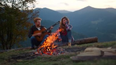 Young couple relax camp in mountains. Hiking hipsters play acoustic guitar at campfire summer mountains. Resting tourists listen perform music outdoors on chill weekend. Vacation recreation concept.