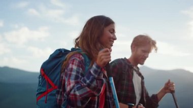 Closeup smiling couple backpack in summer mountains. Happy hikers walk nature outside. Two travelers going trekking poles on tourism destination. Young hipsters talk on sunny day. Recreation concept.