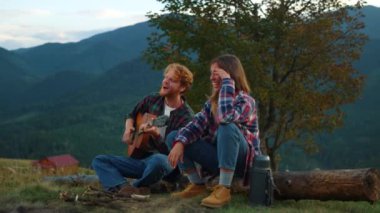 Cheerful couple enjoy nature at evening bonfire closeup. Young travelers play guitar in summer mountains. Smiling campers spend time together on vacation. Family holiday relax leisure travel concept.