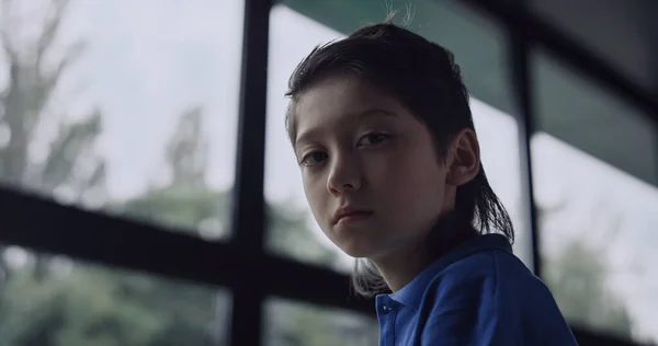 Depressed little boy watching out window feeling loneliness in school close up. Lonely preteen pupil looking camera unhappily sitting empty modern hall. Sad teenager posing alone. Teen crisis concept.