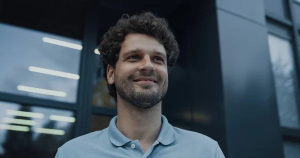 Closeup smiling man teacher standing at school entrance looking distance. Joyful young educational worker welcoming pupils. Attractive curly guy posing at glass door alone. Academic job concept.