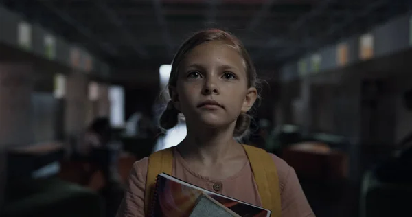 Portrait of tensed little girl walking lively hallway holding books. Focused blonde child going dark corridor on school break alone looking ahead. Stressed frightened pupil worrying about first day.