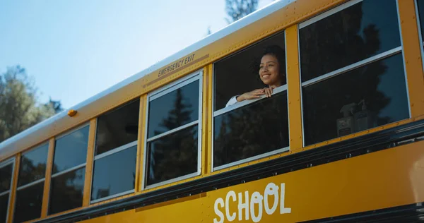 Smiling African American girl looking out schoolbus window in morning. Curly cute elementary age schoolgirl looking in distance alone. Happy schoolchild standing yellow vehicle bus vehicle school.