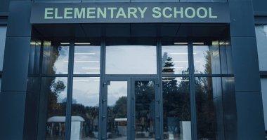 Elementary school exterior with closed glass door. View of window reflecting sky trees. Entrance of new modern campus building on summer vacation without children. Academic year ending concept.
