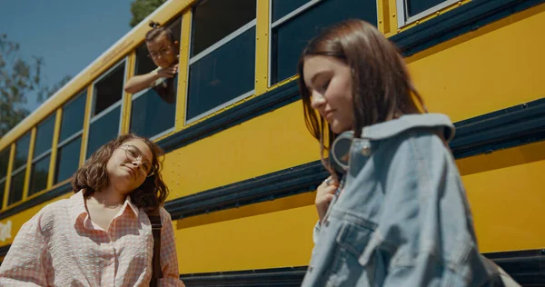 Two cheerful girls talking at yellow school bus. Smiling cute classmates chatting standing near schoolbus in sunlight. Curious schoolboy looking out window observing teen friends. Study concept.