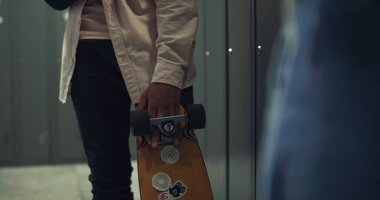 Okul koridorunda üzerinde çıkartmalar olan kaykay tutan çocuk. Dolapların yanında durup arkadaşlarıyla konuşan tanınmamış bir öğrenci. Genç adam alçıya sarılı kolunu kırmış. Hobi konsepti.