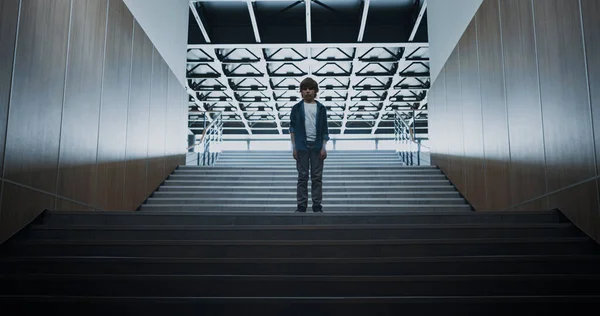 Sad little boy standing alone on modern school staircase. Abandoned schoolboy feel depressed after conflict. Upset preteen guy posing at empty futuristic stairway. Loneliness in childhood concept.