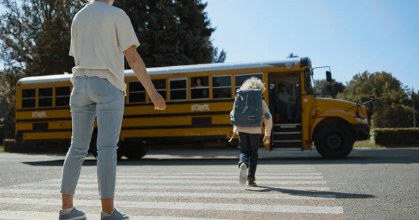 Joyful mom give high five son escorting to schoolbus. Young mother watch child boarding on yellow bus. Happy parent accompany blond boy to school morning time. Schoolboy coming in children transport.