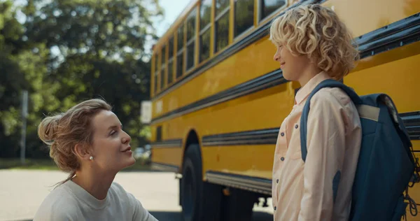 Smiling happy mom chatting with curly son at yellow school bus. Joyful boy wearing backpack standing with mother near academic shuttle. Young woman escorting child to classes. Cheerful family concept.