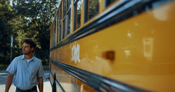 Young school bus driver walking along yellow vehicle. Focused man posing near car. Curly handsome professional worker wearing uniform standing at students transport alone. Chauffeur wait on parking.