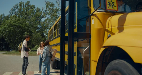 Two preteen classmates leaving yellow school bus open door. Focused serious pupils going out academic shuttle. Diverse cheerful teenagers standing talking together near children vehicle transport.