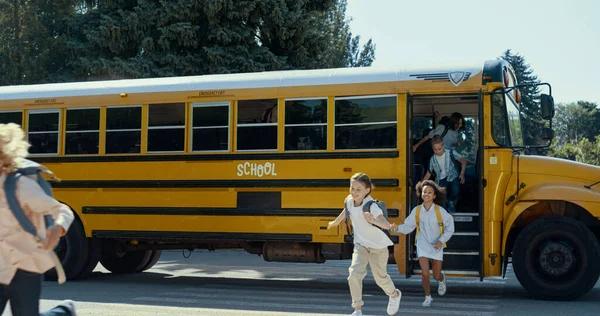 Smiling elementary age students leaving yellow school bus. Cheerful diverse schoolchildren running jumping out of academic shuttle. Man driver watching pupils onboarding. Public education concept.