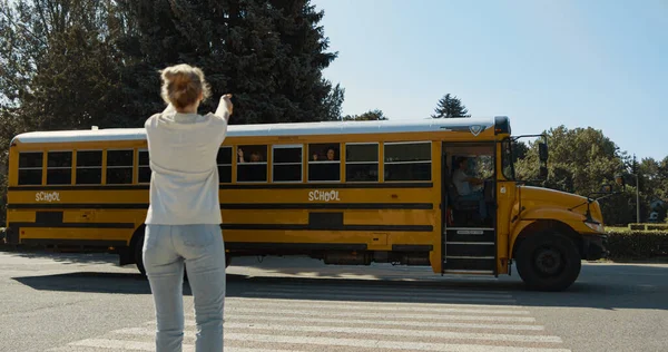 Happy mom standing waving to child sitting yellow schoolbus. Joyful pretty woman send air-kiss to little son summer sunny morning. Caring parent saying goodbye schoolboy before leaving to school.