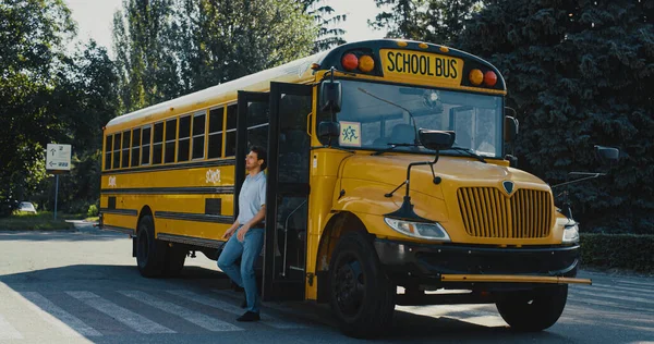 Young man driver coming out schoolbus cabin. Empty school bus standing on parking lot. Professional chauffeur wearing uniform checking equipment alone sunny day. Schoolchildren transportation concept.
