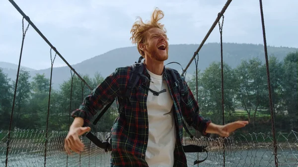 Euphoric hiker jump outdoors on river bridge. Close up joyful backpacker feel happy in mountains. Excited tourist enjoy celebrate success victory. Cheerful ginger traveler on holiday. Active concept