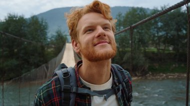 Neşeli turist suratı nehir köprüsünde yürümekten zevk alıyor. Kızıl saçlı gezgin dağ yürüyüşü portresi. Gülümseyen neşeli sırt çantalı yeşil manzaraya bir bak. Yazın yürüyüşe çıkan mutlu adam. Macera konsepti.