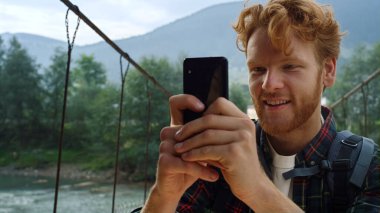 Neşeli yürüyüşçü cep telefonuyla video çekiyor. Zencefilli sırt çantasıyla Nehir Köprüsü 'nde seyahat blogunun keyfini çıkar. Dağlarda gezici cihaz kullanarak fotoğraf çeken mutlu bir turist. Seyahat modern tatil konsepti.