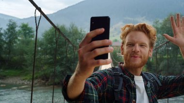 Dağlarda cep telefonu kamerasıyla heyecanlı bir yürüyüşçü görüntüsü. Duygusal hipster el sallamasını cep telefonuyla selamlayın. Doğa manzarasında blog yazan mutlu kızıl adam. Seyahat tatili kavramı