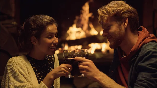 Couple celebrate evening date by fireplace. Close up relaxed lovers drink red wine on vacation. Happy family clink glasses on honeymoon. Husband wife enjoy alcohol on anniversary. Toast love concept.