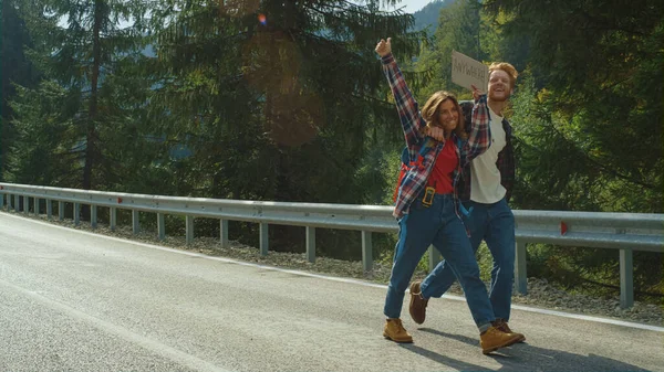 Excited friends hiking mountain road. Two backpackers hold text banner on hitchhike. Laughing tourists show thumbs up on vacation trip. Happy couple enjoy hug on honeymoon adventure. Travel concept.