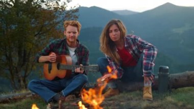 Closeup playful couple spend holiday sit bonfire. Young lovers play acoustic guitar music in mountains. Two joyful campers have fun at romantic nature date outdoors. Travel lifestyle leisure concept.