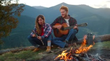 Relaxed tourists enjoy bonfire spend evening in mountains. Close up happy couple camp outdoors on sunset nature. Romantic lovers play guitar on holiday weekend. Family activity together concept.