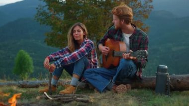 Closeup couple travelers relax together on mountains trip. Lovely campers enjoy romantic music. Affectionate lovers look each other on evening bonfire. Chill tourists play guitar. Leisure concept.