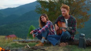 Positive couple date outdoors in mountains. Close up two lovers play guitar by campfire. Carefree travelers relax on activity. Resting campers enjoy music on romantic evening picnic. Relax concept.