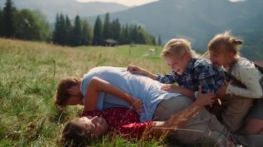 Joyful family lying green grass piled on each other sunny day. Playful parents laughing fooling with children on beautiful mountain hill. Happy couple playing with kids on nature summer holiday. 