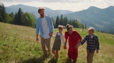 Happy family enjoying walk together in mountains summer holiday. Relaxed parents pointing fingers on beautiful nature views hiking with children. Smiling couple with kids holding hands on green hill.