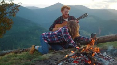 Camping tourists enjoy picnic on mountains hike. Young couple relax by bonfire close up. Joyful family play guitar music on family weekend. Smiling travelers spend holiday on nature. Outdoors concept.