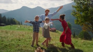 Playful family having fun summer vacation in mountains. Happy parents enjoying active funny games on beautiful green hill. Joyful woman fooling touching children playing on grass with redhair man.