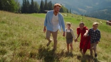 Friendly family walking on mountain slope sunny day. Happy parents holding children hands going to top green hill. Positive mother pointing finger on beautiful nature landscape. Vacation together.