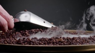 Kavrulmuş kahve çekirdekleri kepçeyle kepçeyle kaplanıyor. Kimliği belirsiz bir adam elinde kepçeyle kızartma makinesinde aromatik tahıl karıştırıyor. Kahverengi kahve çekirdeklerinin üzerine ağır çekimde hafif buhar yükseliyor..