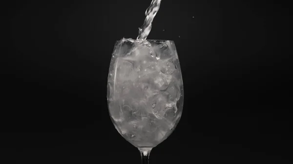 Mineral water pouring ice glass closeup. Jet of fizzy drink filling wineglass with ice cubes. Sparkling bubbled aqua with splashes black background. Preparing cocktails and beverages concept