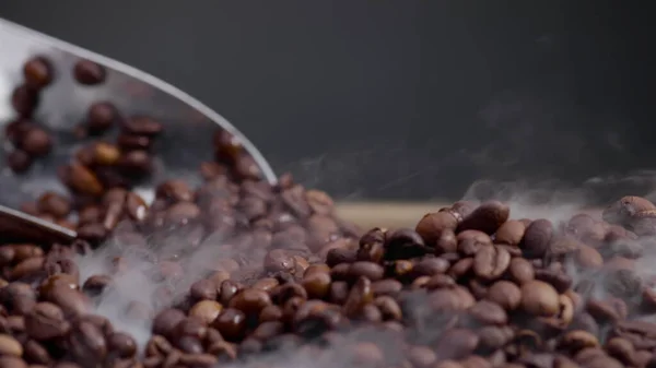 Metal ladle scooping up roasted coffee beans from burlap sack close up. Unknown person mixed aromatic seeds using shovel in super slow motion. Light smoke coming over fresh brown coffee grains.