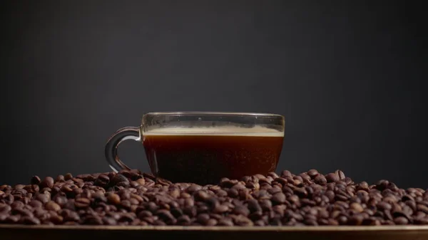 Fragrant coffee drops falling in full espresso cup close up. Transparent mug standing on heap roasted aromatic seeds. Aromatic energetic morning beverage placed on pile brown grains. Arabica concept.