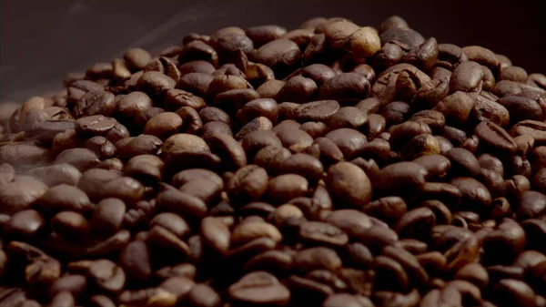 Macro view coffee seeds isolated on dark background. Close up light vapor rising on roasted fresh grains heap in super slow motion. Beautiful coffee beans roasting steaming on pile. Caffeine beverage.