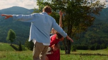 Playful family running with outscretched arms on green hill. Joyful parents playing with children outdoor games on meadow. Happy couple with kids having fun in mountains enjoying summer vacation.